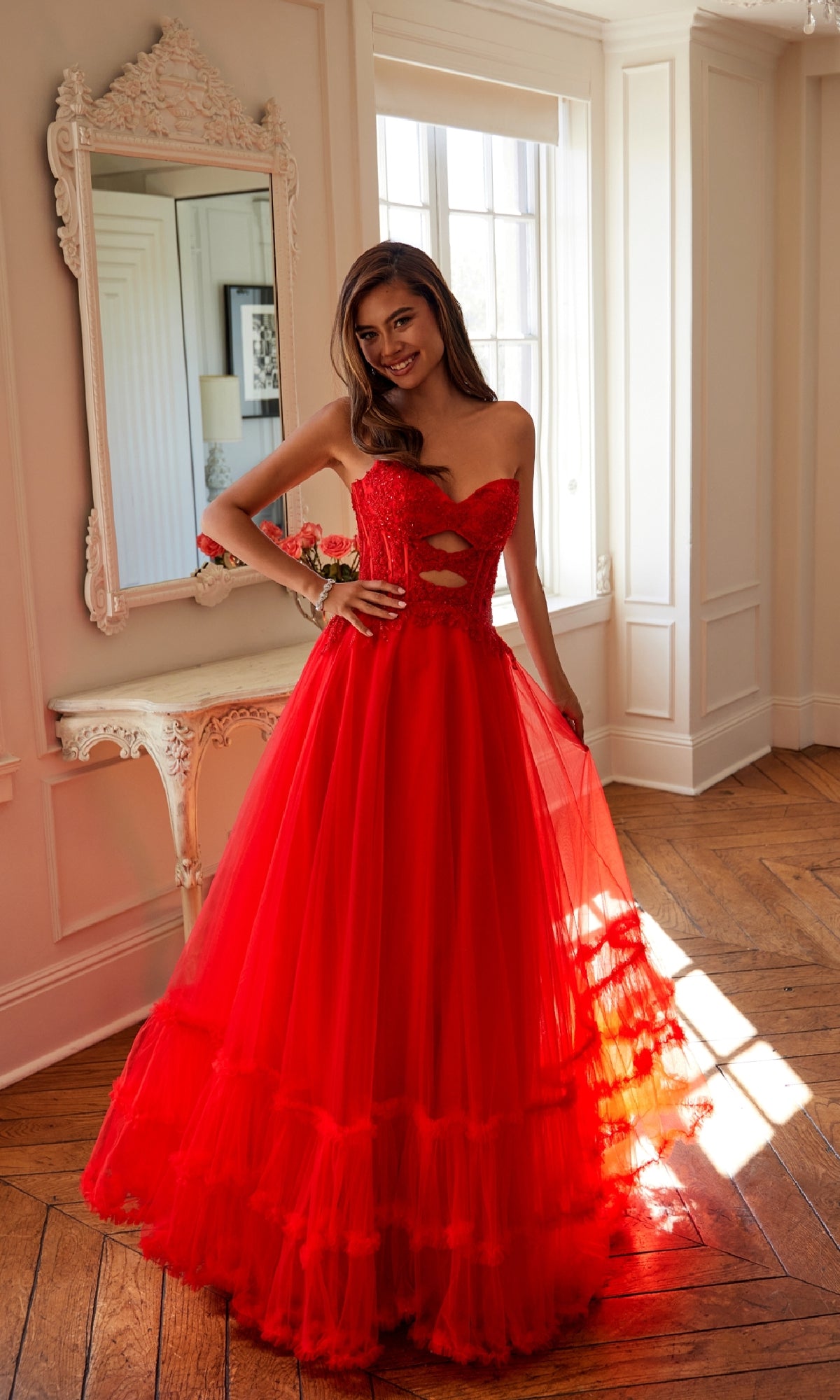 A woman poses indoors near a window and white ornate mirror, wearing the Strapless Long Prom Ball Gown: Juliet JT2561S featuring an embroidered-lace bodice and tulle skirt.
